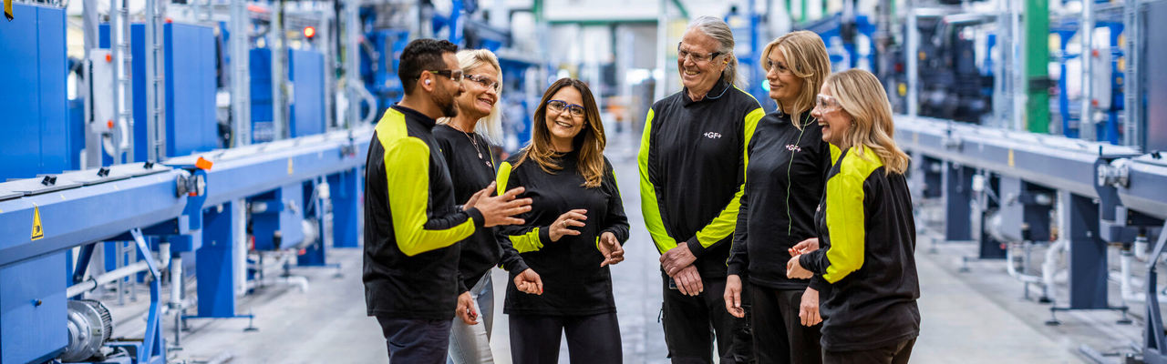 Sechs GF Mitarbeitende stehen gemeinsam in einer modernen Industrieumgebung und tauschen sich aus.