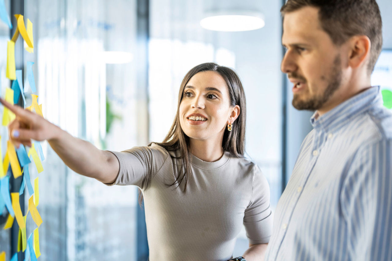 Two employees brainstorming in front of a glass wall covered with sticky notes.